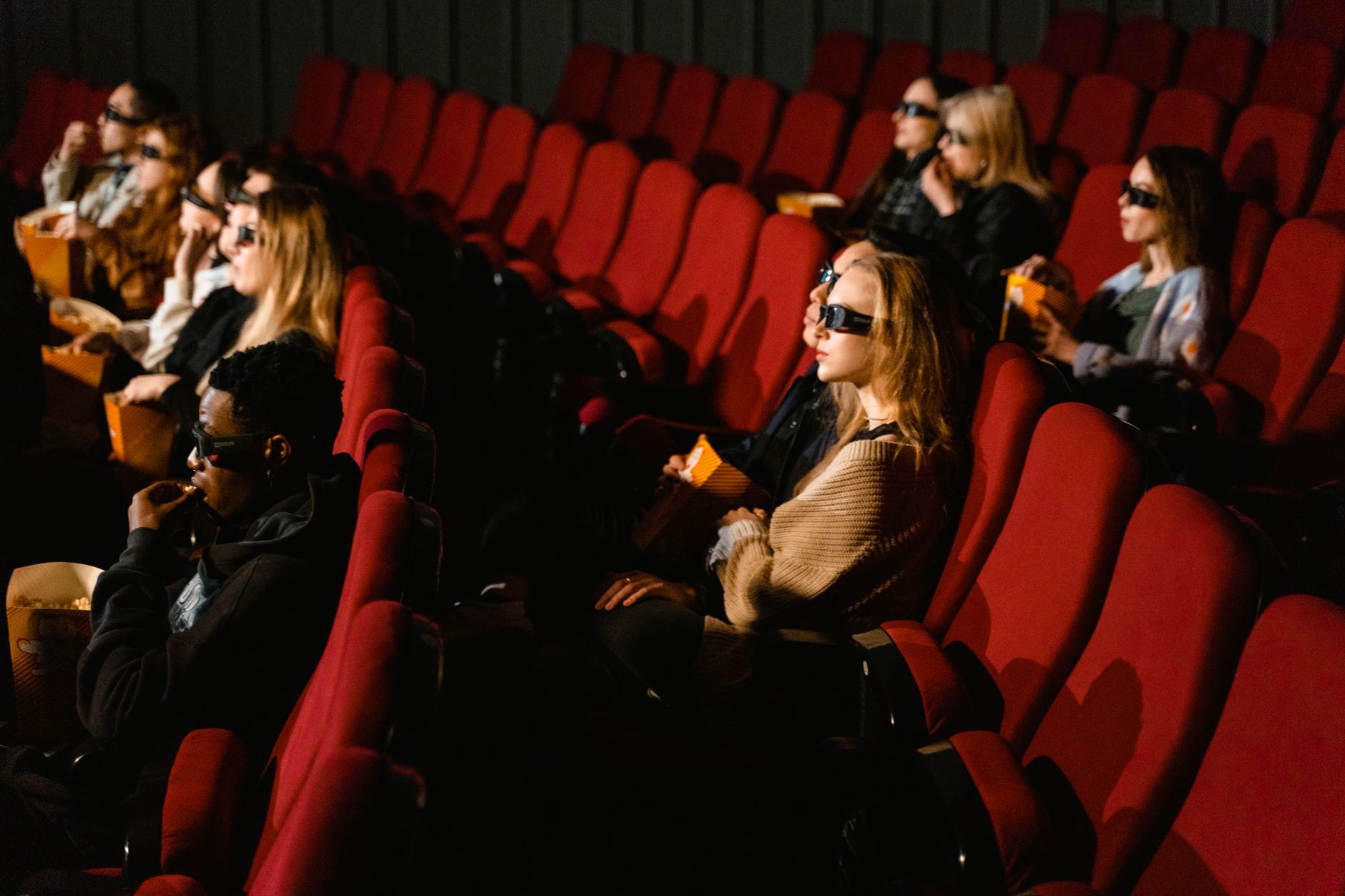 Gens portant des lunettes 3D et mangeant du pop-corn dans un cinéma avec des sièges rouges.