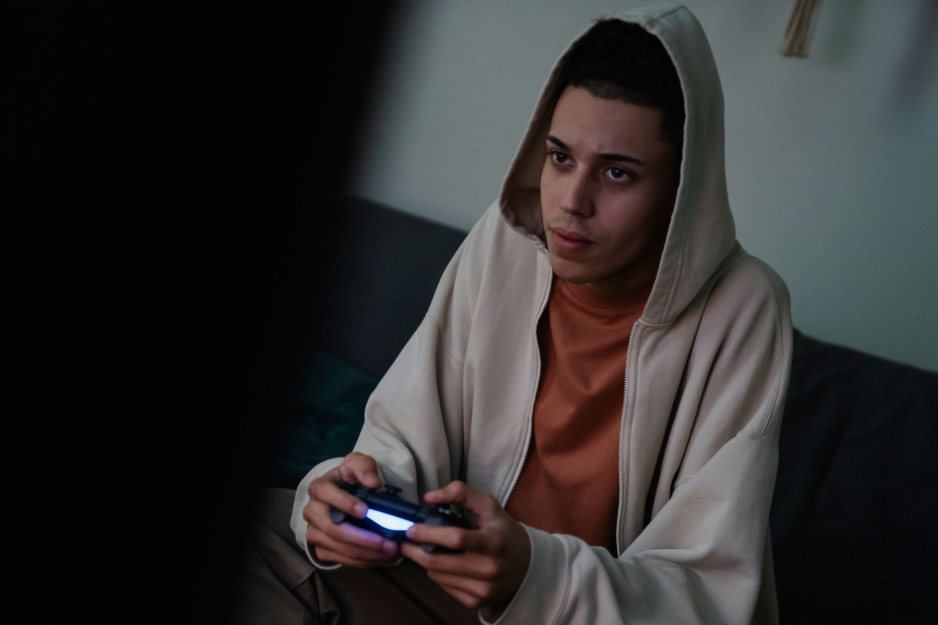 Jeune homme en sweat à capuche beige assis sur un canapé, une manette de jeu à la main, dans une pièce sombre.