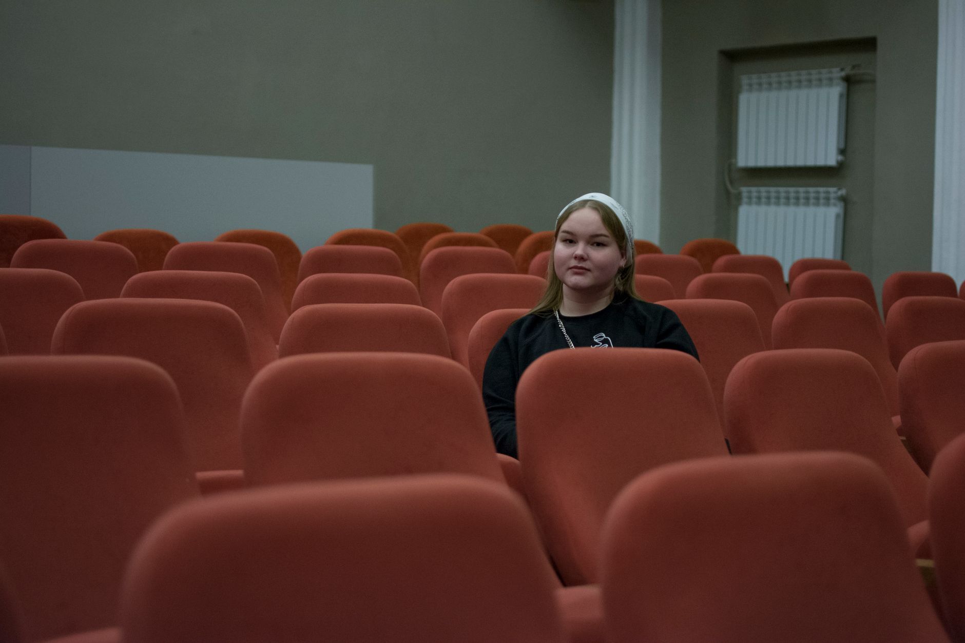 Jeune personne assise seule parmi des rangées de fauteuils rouges dans une salle de projection vide.