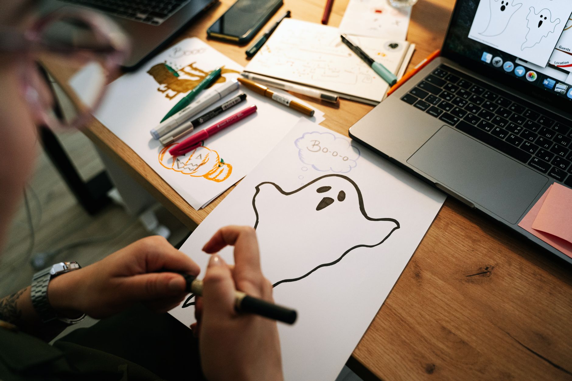 Personne dessinant un fantôme au feutre noir sur une feuille, entourée de croquis, de feutres et d’un ordinateur portable sur un bureau en bois.