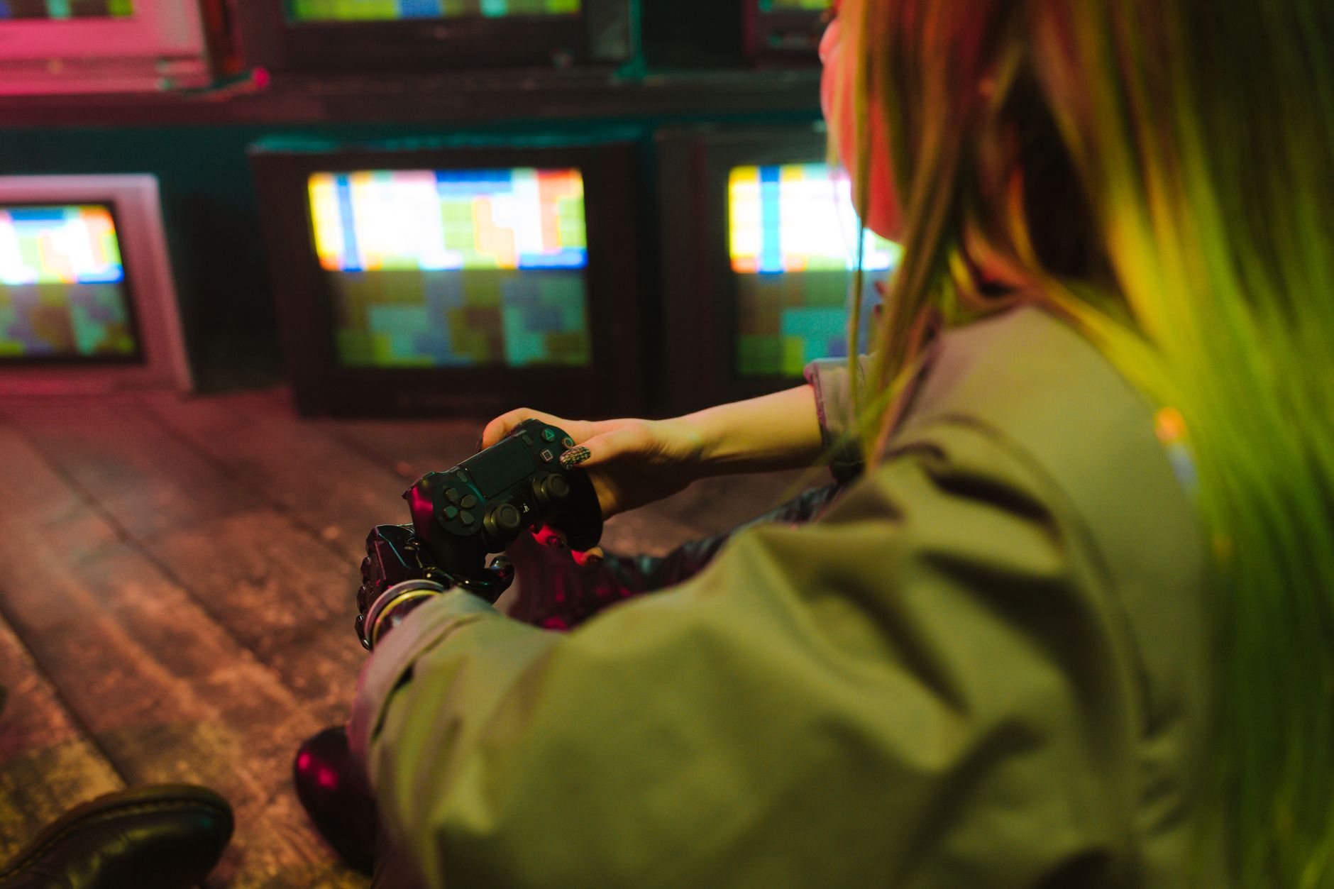 Personne vue de dos tenant une manette de jeu noire devant des téléviseurs anciens aux écrans colorés, dans une pièce sombre éclairée de rose et de vert.