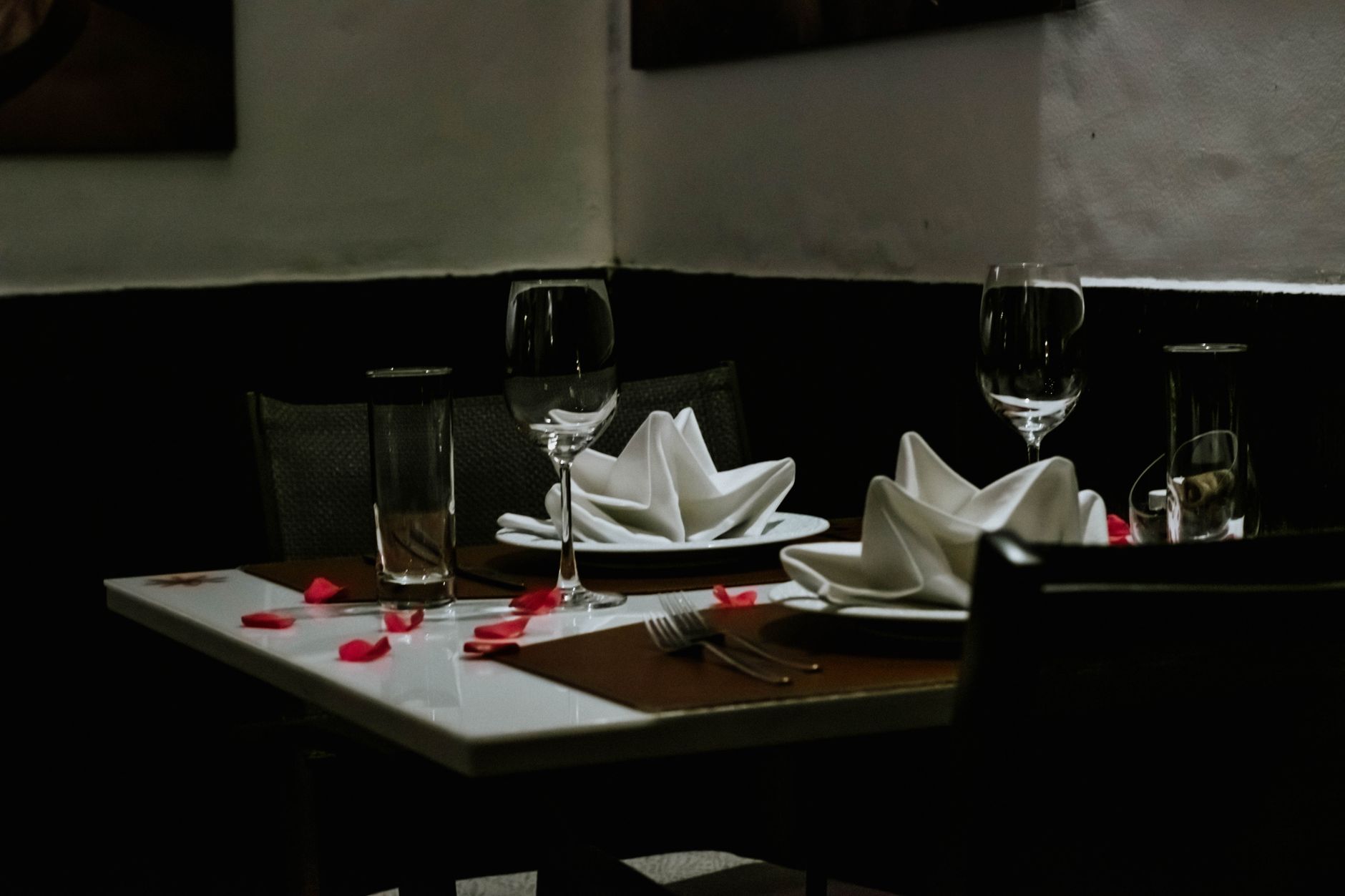 Table de restaurant dressée pour deux, avec serviettes blanches pliées, verres à pied et pétales de rose rouges, dans une salle sombre.