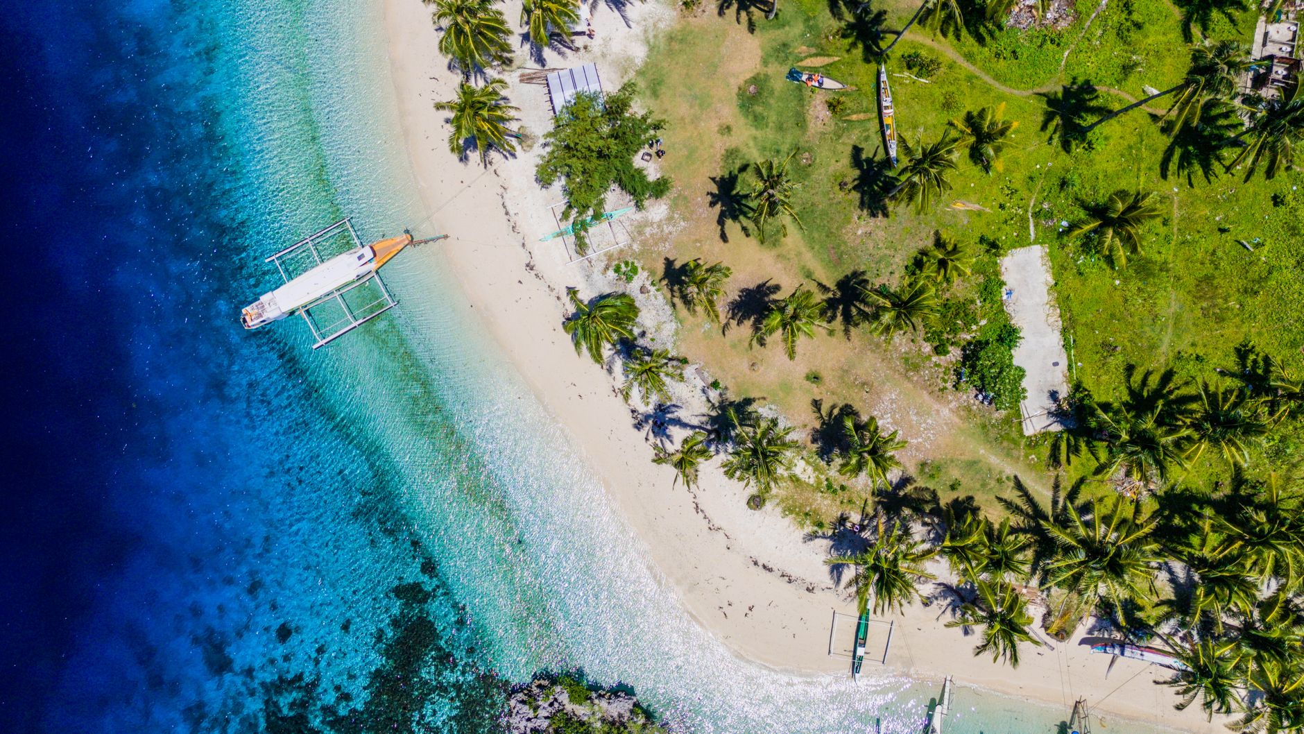 Vue aérienne d’une plage tropicale avec un bateau à balancier dans une eau turquoise, bordée de palmiers.
